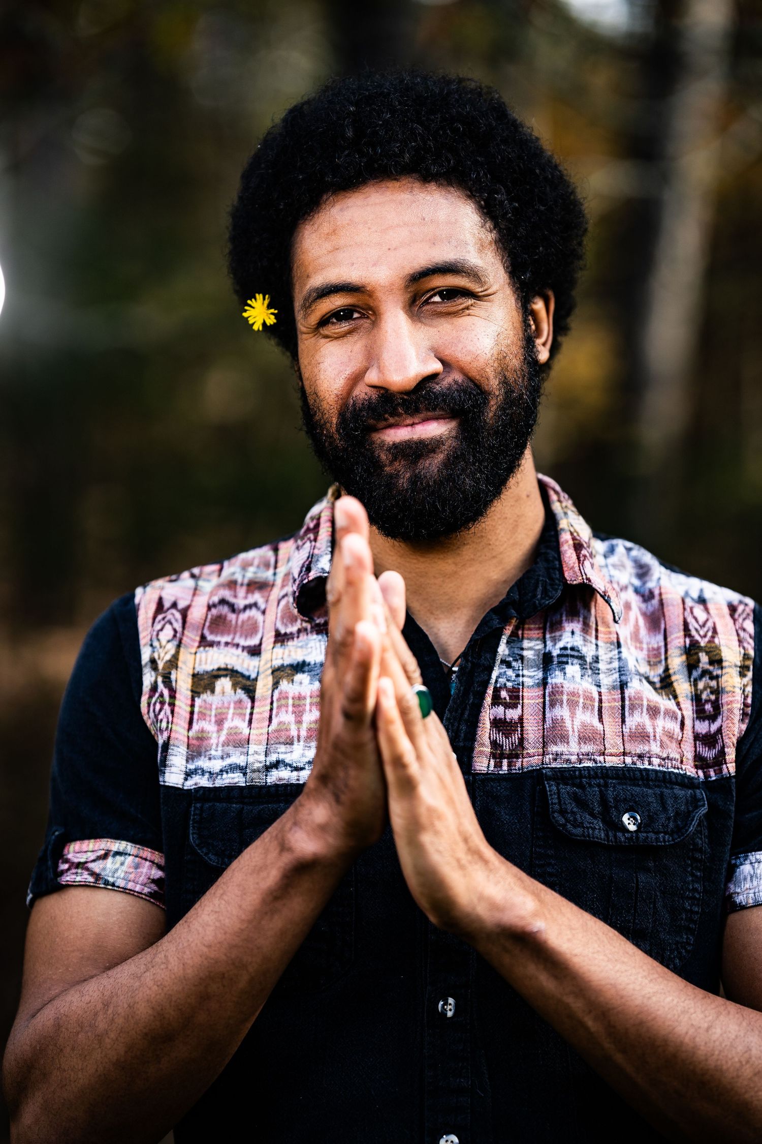 Smiling bearded Black man with afro and yellow flower, hands in prayer pose, wearing colorful patterned vest outdoors.