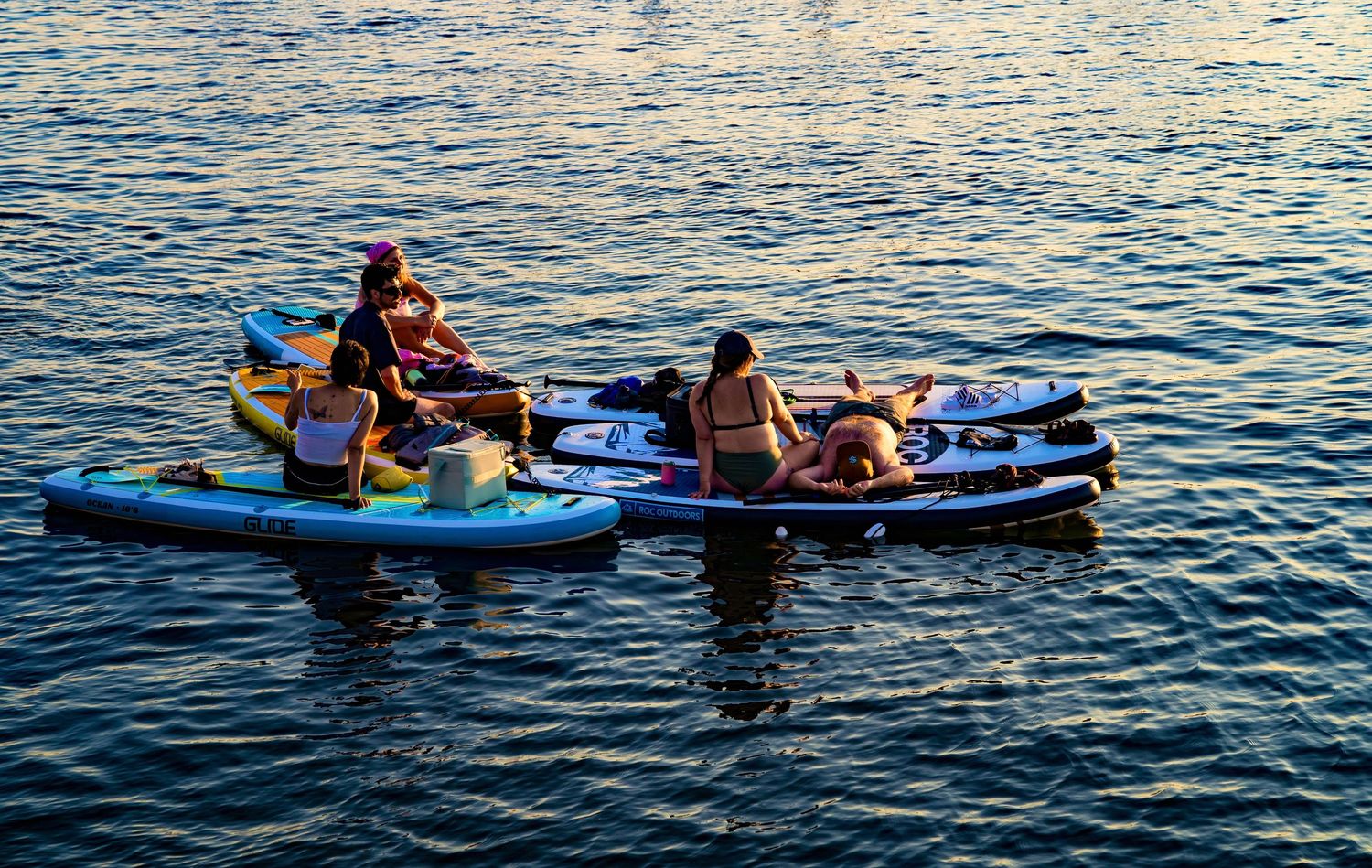 Group of people sitting on paddleboards floating on calm blue water during sunset.