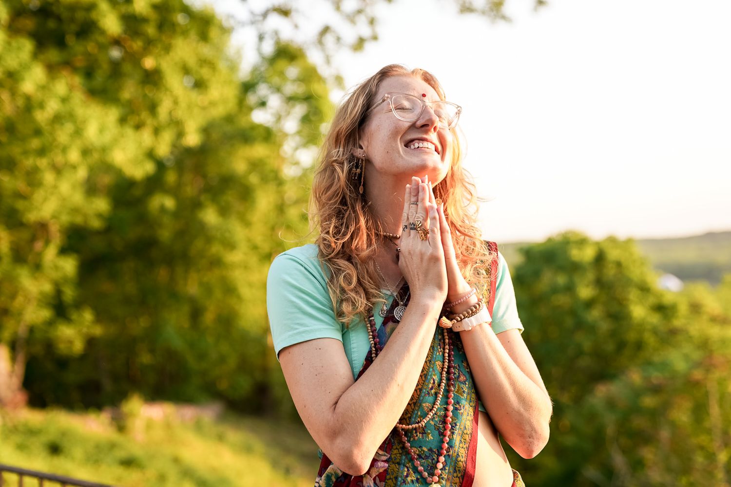 Woman with curly hair in prayer pose smiling outdoors, surrounded by lush green trees in warm sunlight.