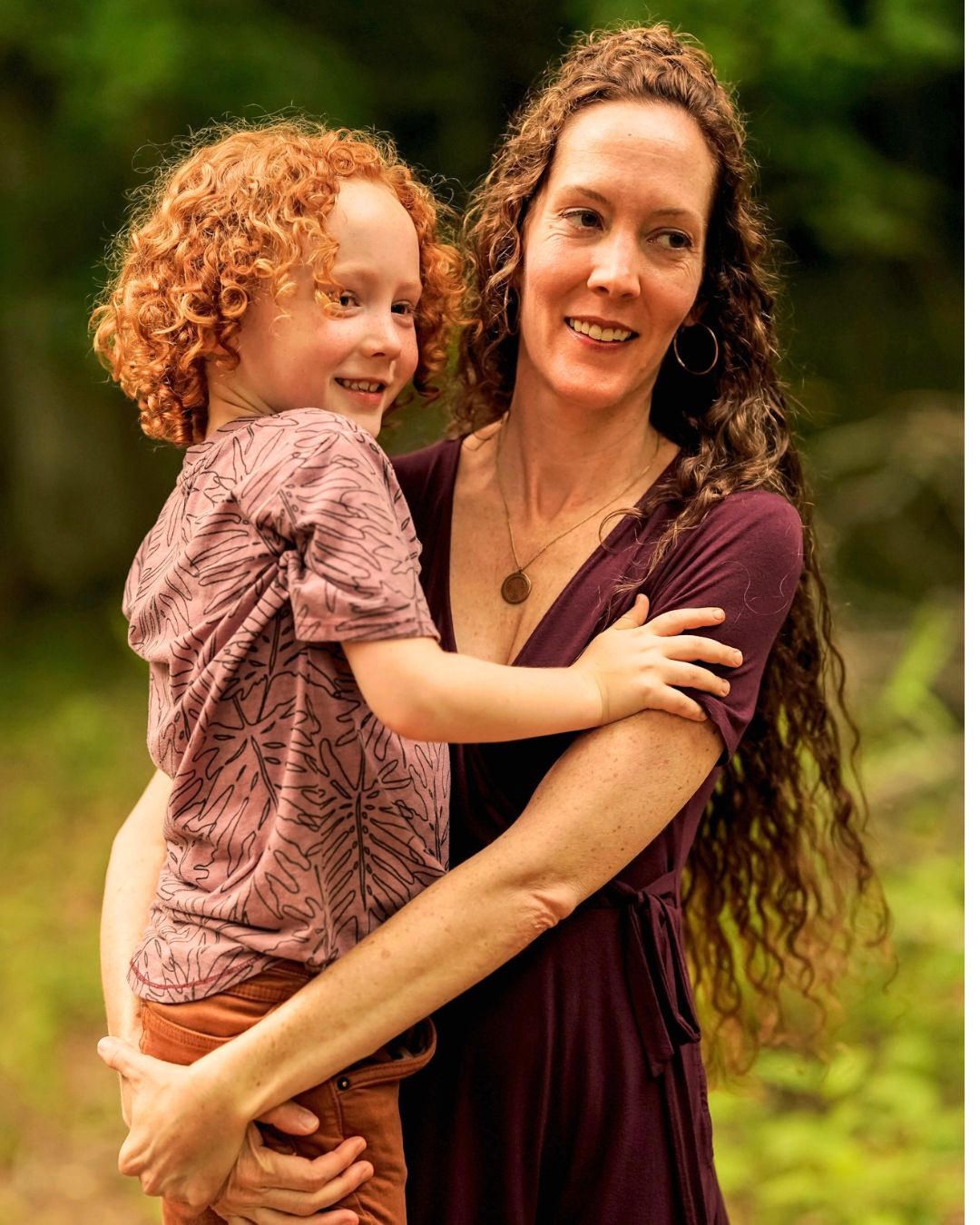 A woman with curly hair holds a young redheaded child outdoors, both smiling warmly in a green natural setting.