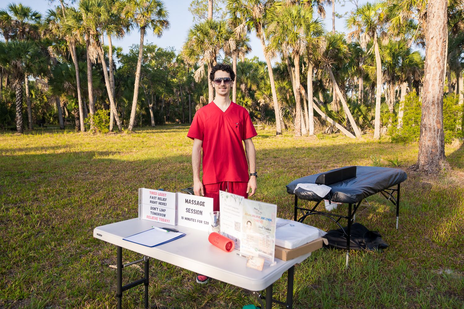 A man wearing a red shirt standing at his booth as a massage therapist, positioned next to a massage table.