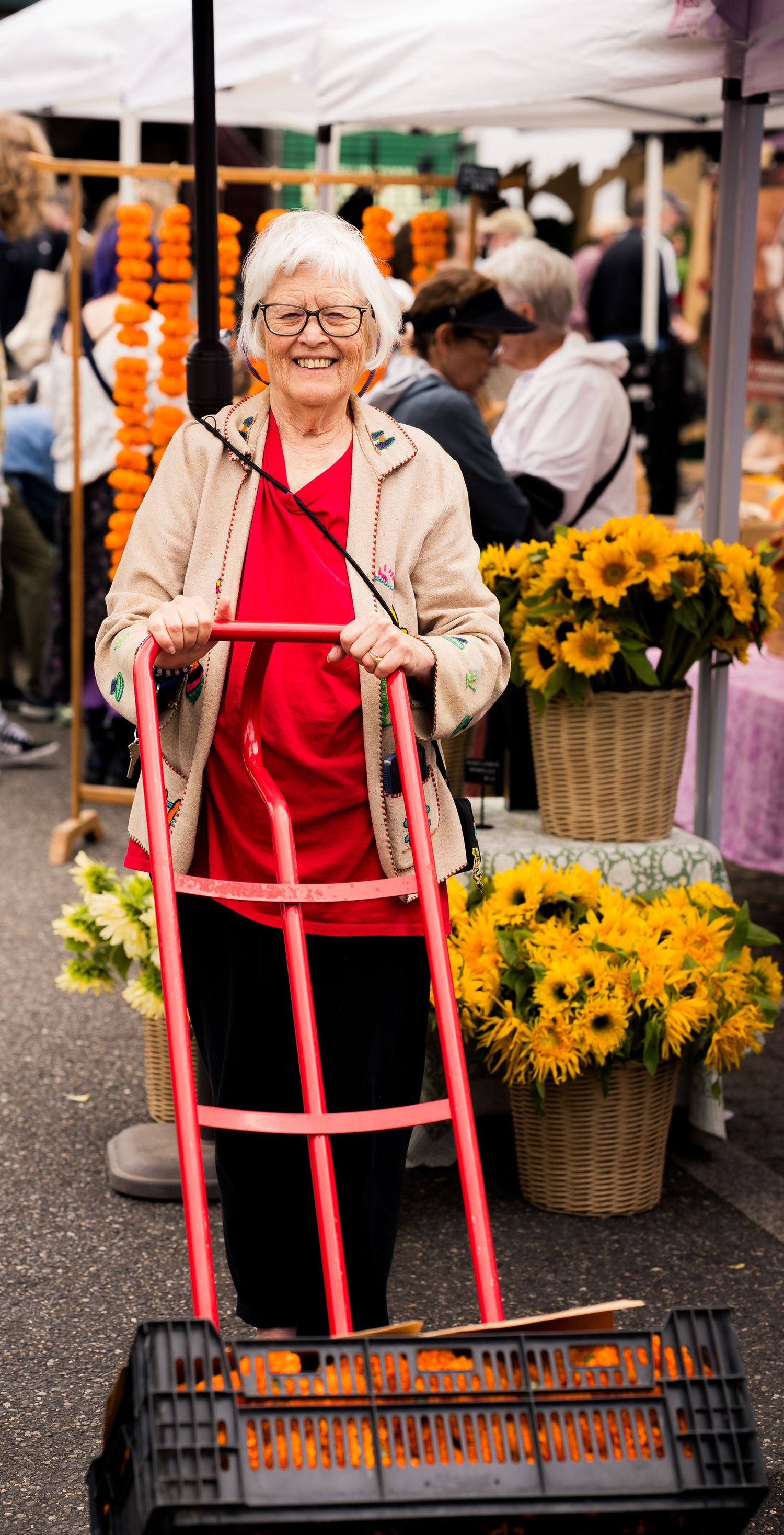 Smiling elderly woman with walker at outdoor flower market, surrounded by sunflowers and marigold garlands.