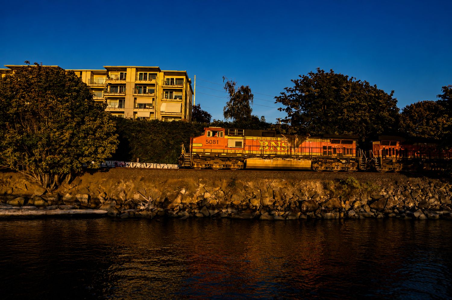 Orange BNSF freight train traveling along a rocky waterfront beside lush trees and a modern apartment building at sunset.