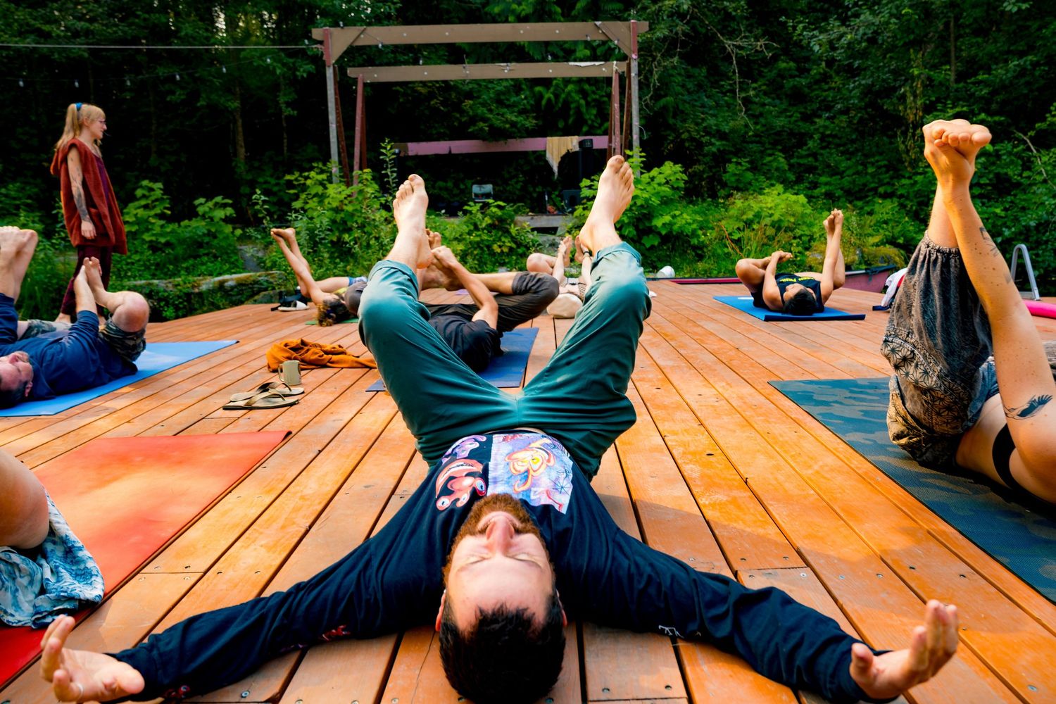 Group yoga class on outdoor wooden deck surrounded by lush green forest, participants in reclined poses on colorful mats.