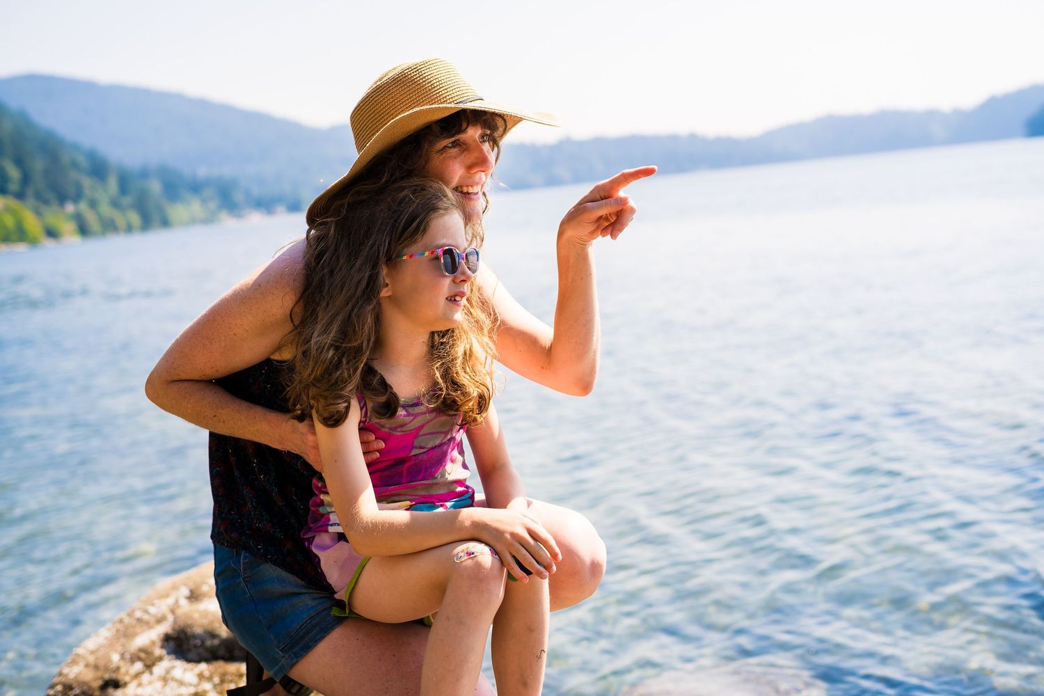 Woman and girl sitting on rocks by a lake, pointing at the distance, wearing sunglasses and a straw hat.