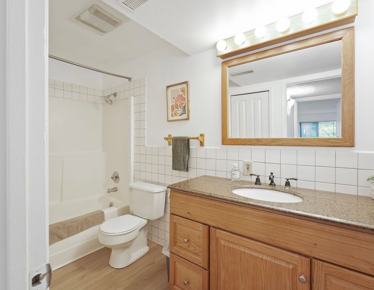 Clean full bathroom featuring oak vanity with granite countertop, white tub-shower combo, toilet, and large mirror with lighting.