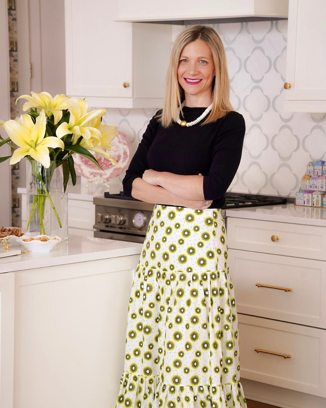 Woman in floral skirt and black top posing in elegant white kitchen with yellow lilies.