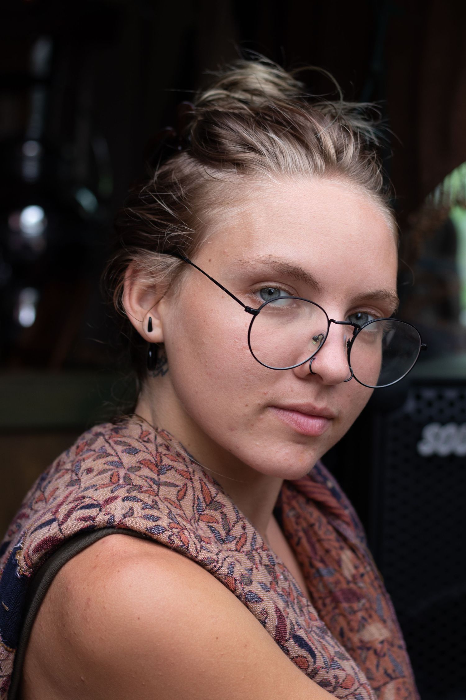 A blonde girl wearing glasses, looking slightly toward the camera with a gentle expression.