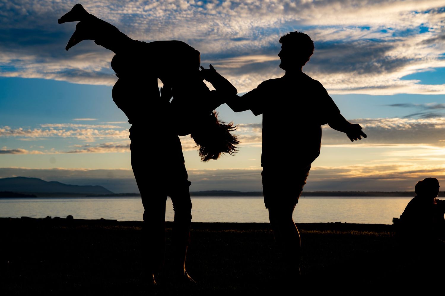 Silhouettes of people playing at sunset near a lake with a dramatic cloudy sky in the background.