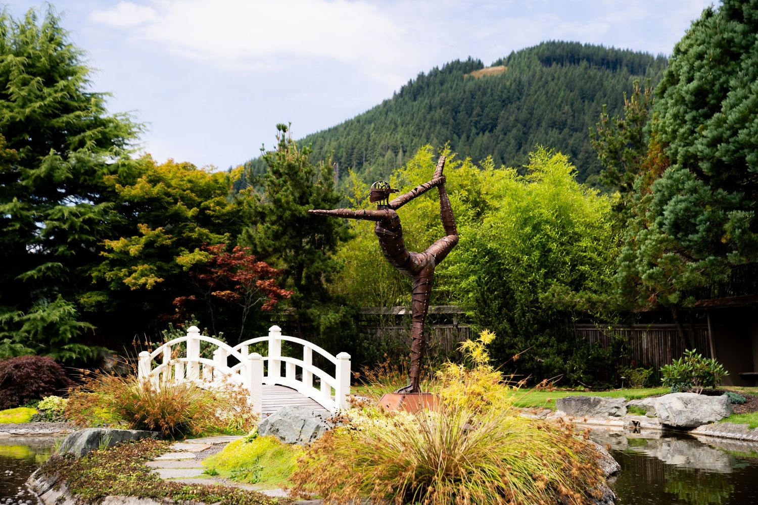 Serene Japanese garden with white arched bridge, sculptural tree, lush greenery, and forested mountain backdrop.