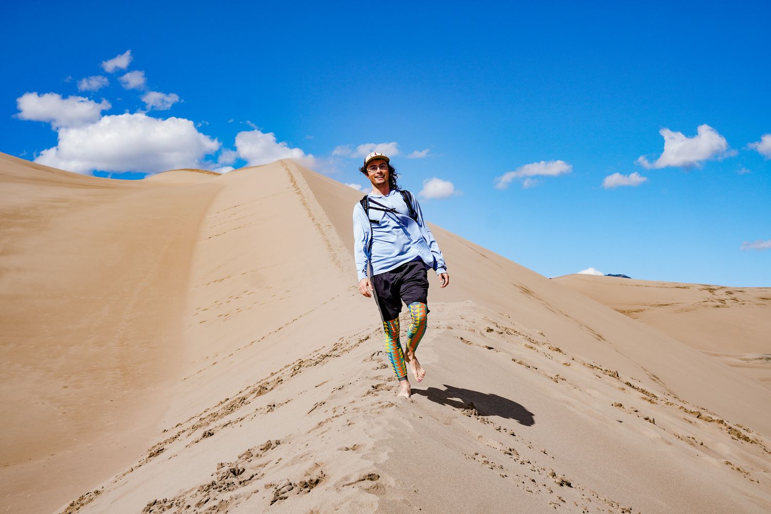 A hiker walks down a sandy desert dune under a bright blue sky with scattered clouds.