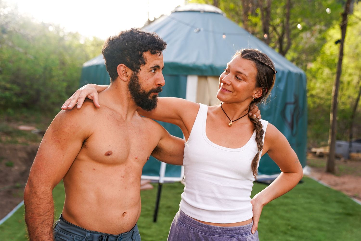 Smiling couple stands together near a yurt in a lush green outdoor setting on a sunny day.