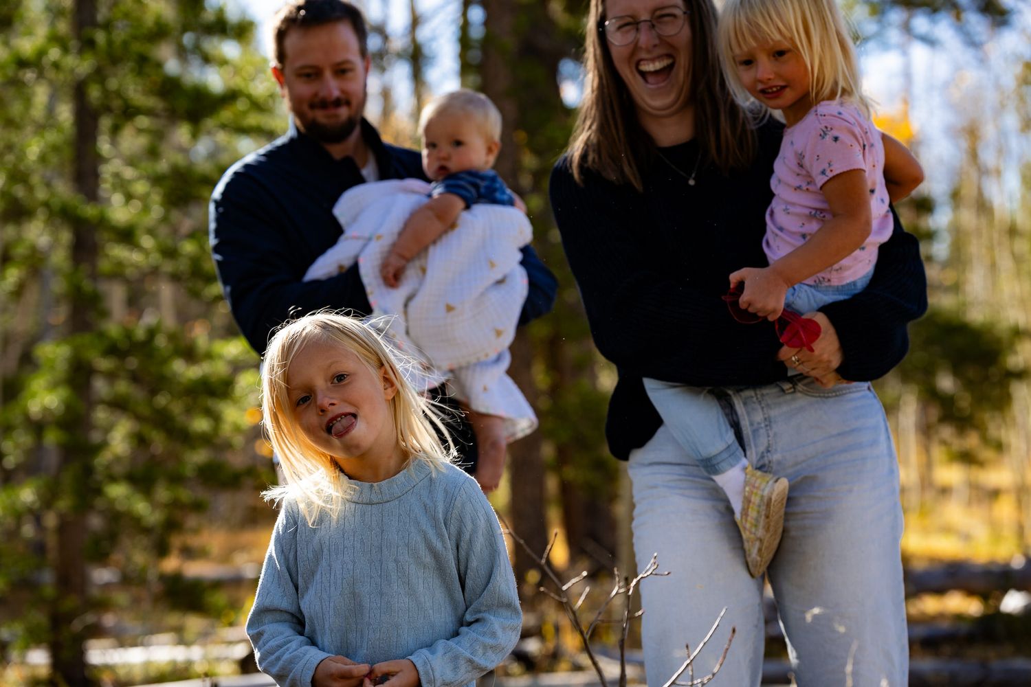 Happy family with three young children laughing outdoors in sunny pine forest.