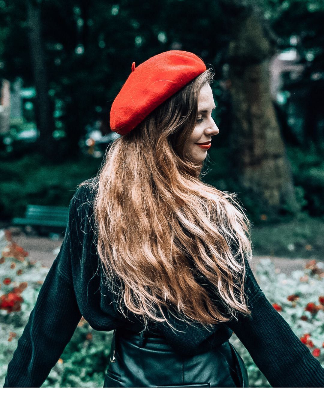 Woman in red beret and black outfit with long wavy hair standing in a lush green park.