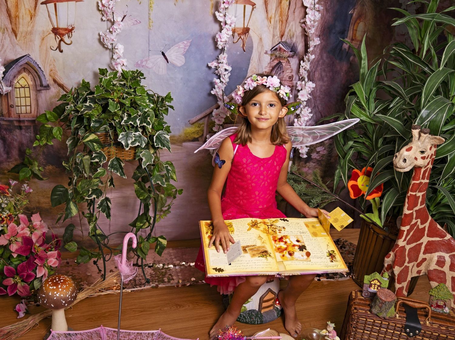 A young girl in a pink dress and fairy wings holds a large book in a magical fairy tale garden setting.