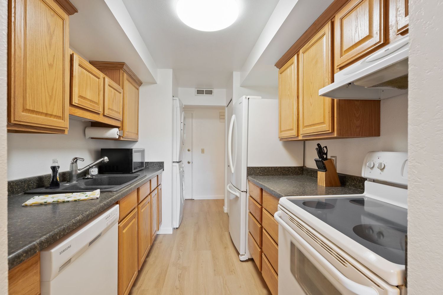 Modern galley kitchen with oak cabinets, dark granite countertops, white appliances, and light hardwood floors.