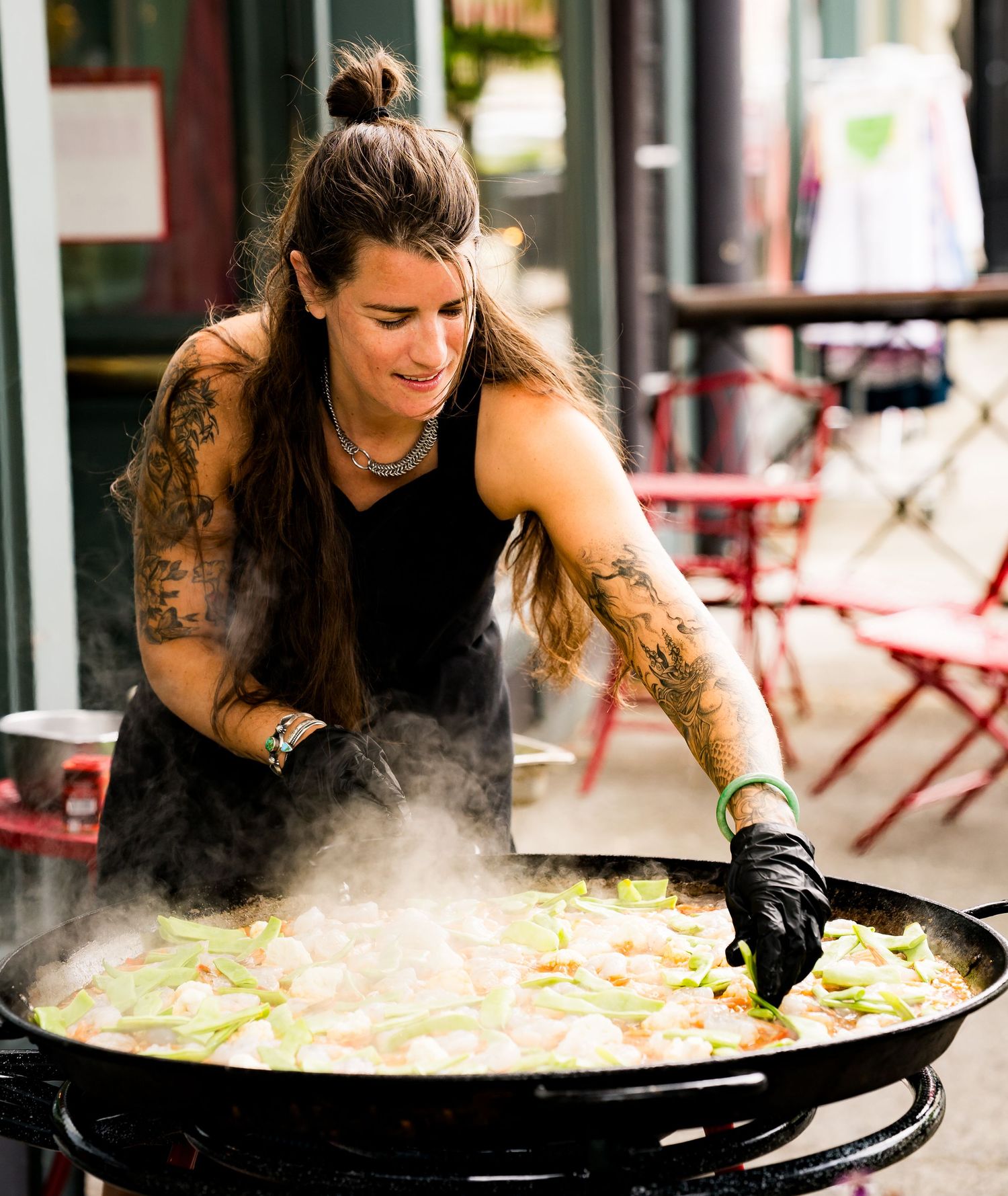 Tattooed woman wearing black gloves stirs steaming vegetables in a large outdoor paella pan at a street food event.