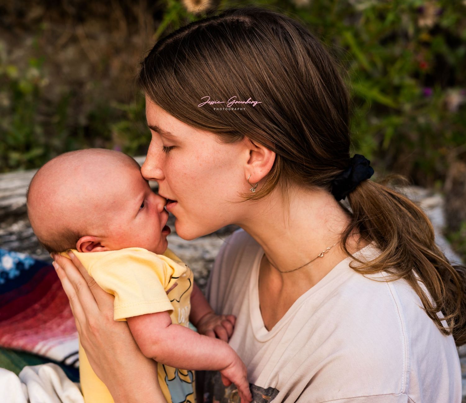 A young mother tenderly kisses her newborn baby outdoors, sharing a sweet bonding moment in natural light.