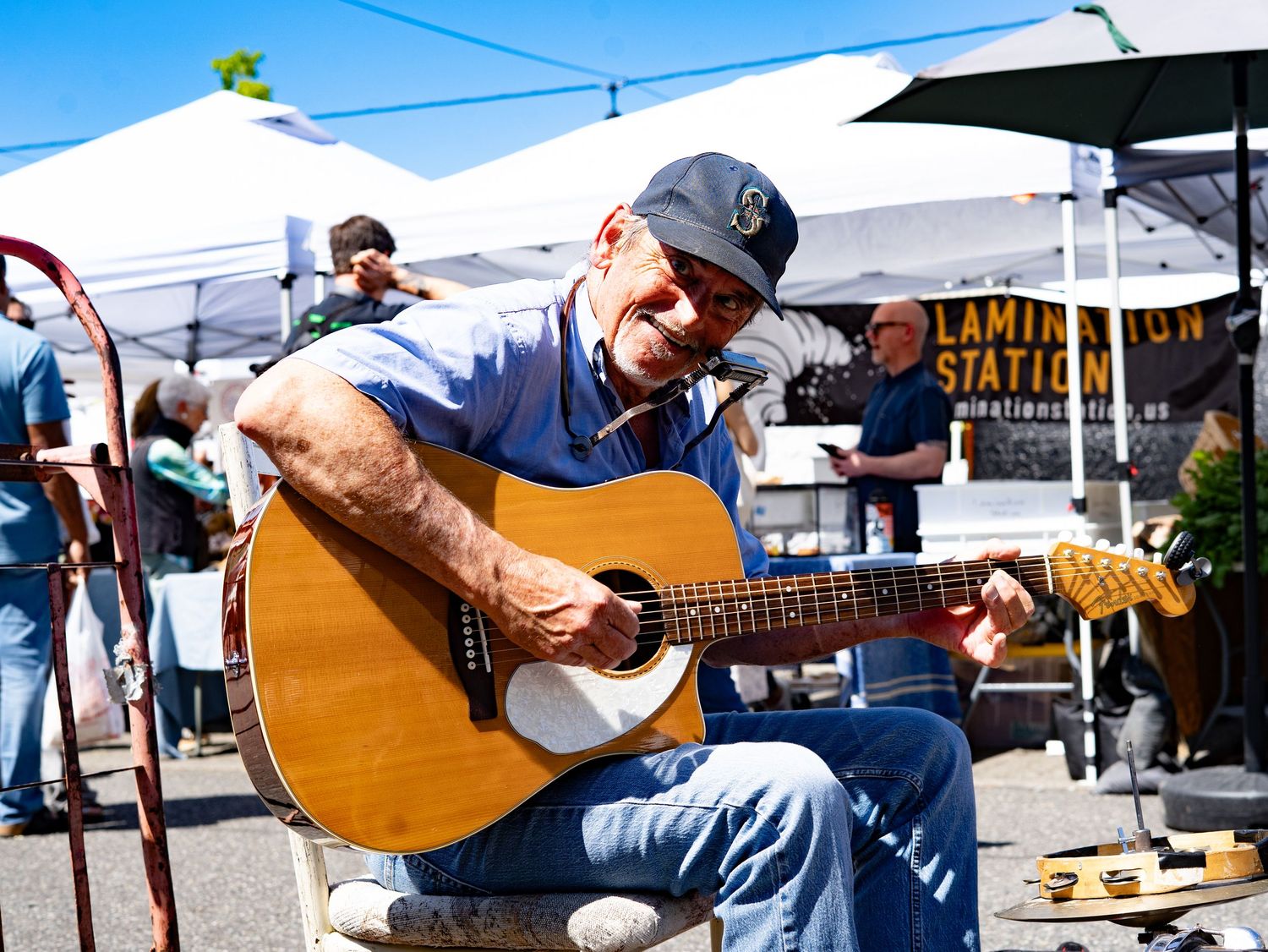 Elderly man playing acoustic guitar outdoors at a market, smiling joyfully while seated under white tents on a sunny day.
