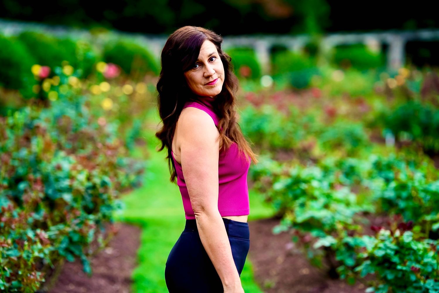 A woman in a pink top poses outdoors in a lush green garden setting during a portrait photoshoot.
