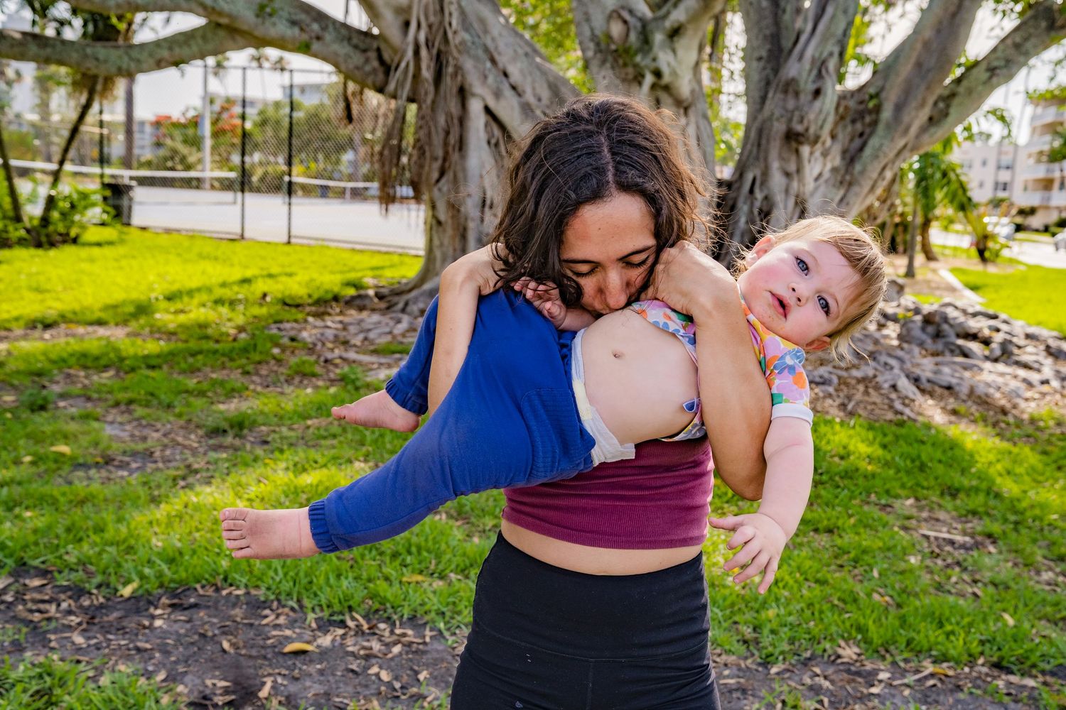 A photo of the mom with a pink shirt kissing the babies belly with a tree and grass behind them.