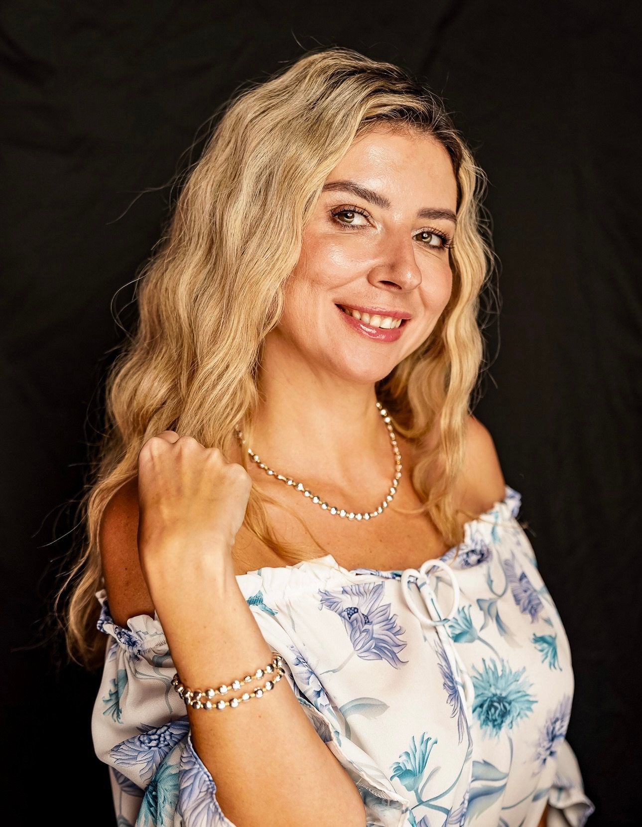 A picture of a woman with blonde hair and a top showing her shoulders, she is smiling and looking at the camera in a studio, black backdrop.