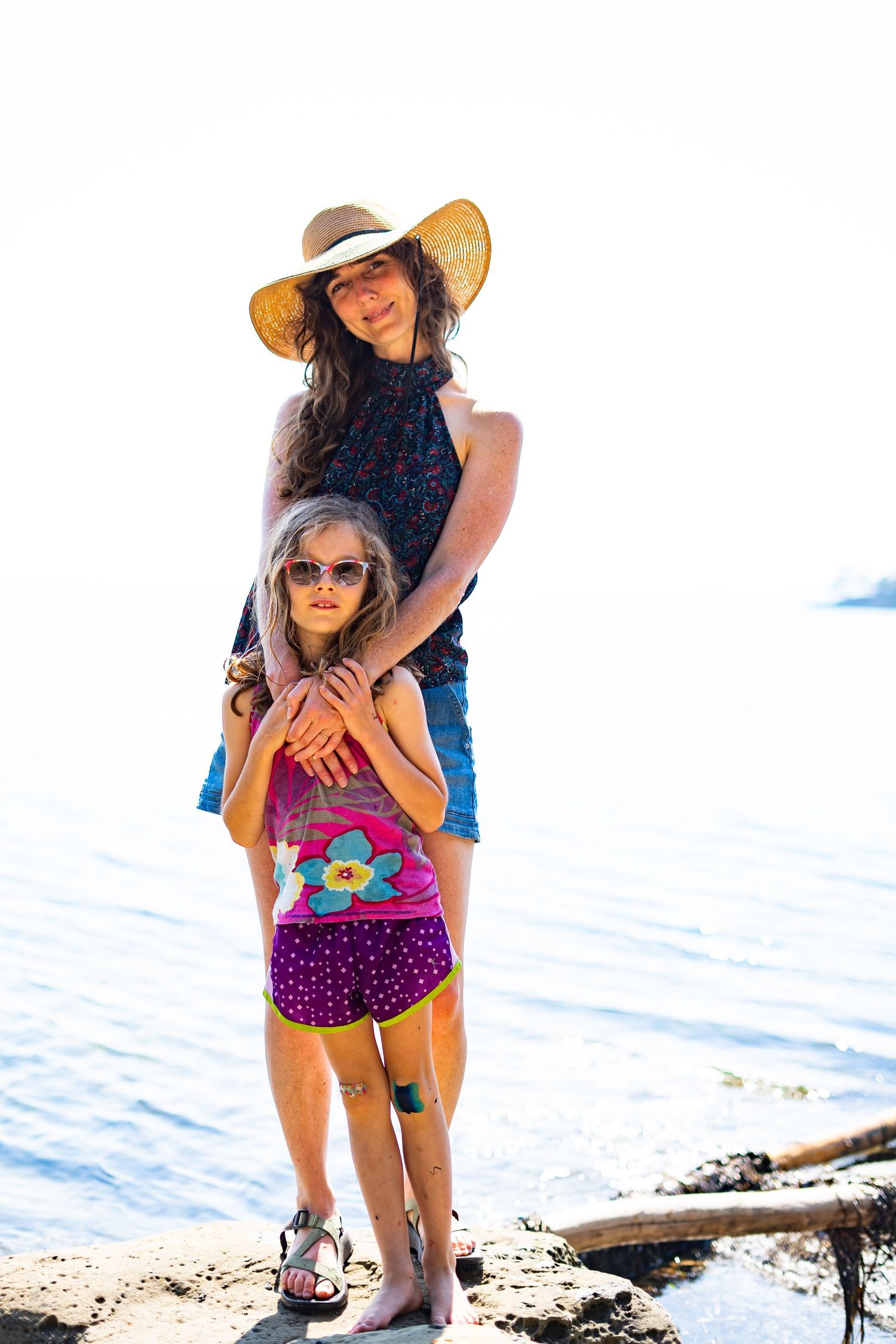 Woman in straw hat embracing young girl in sunglasses on rocky beach shore on a sunny day.