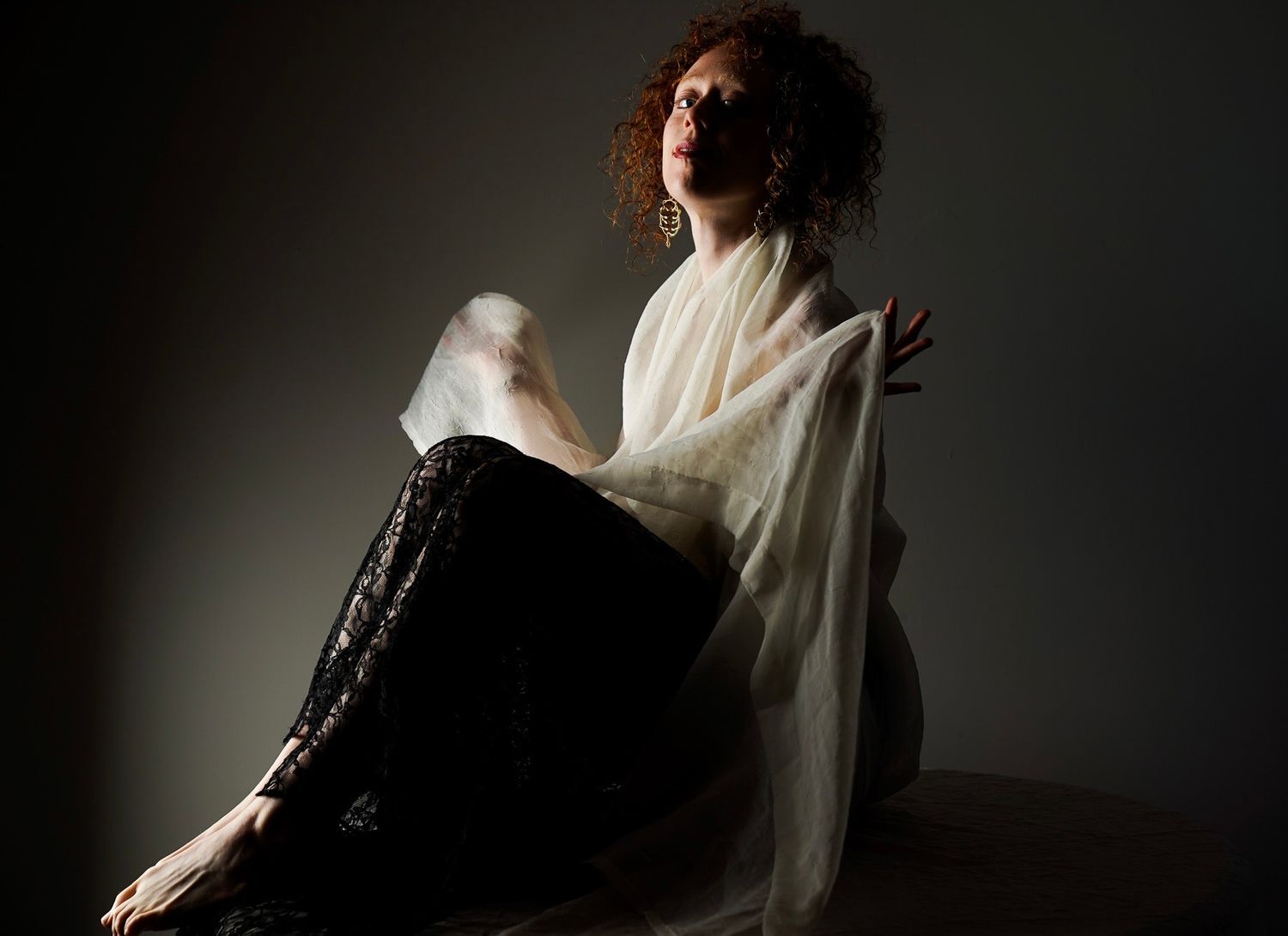 A woman with curly red hair sits gracefully draped in a sheer white fabric against a dark studio background.
