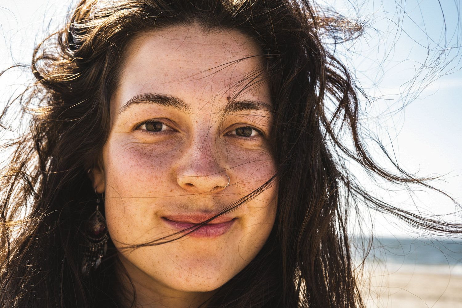 Young woman with dark wavy hair blowing in the wind smiles at the beach on a sunny day.