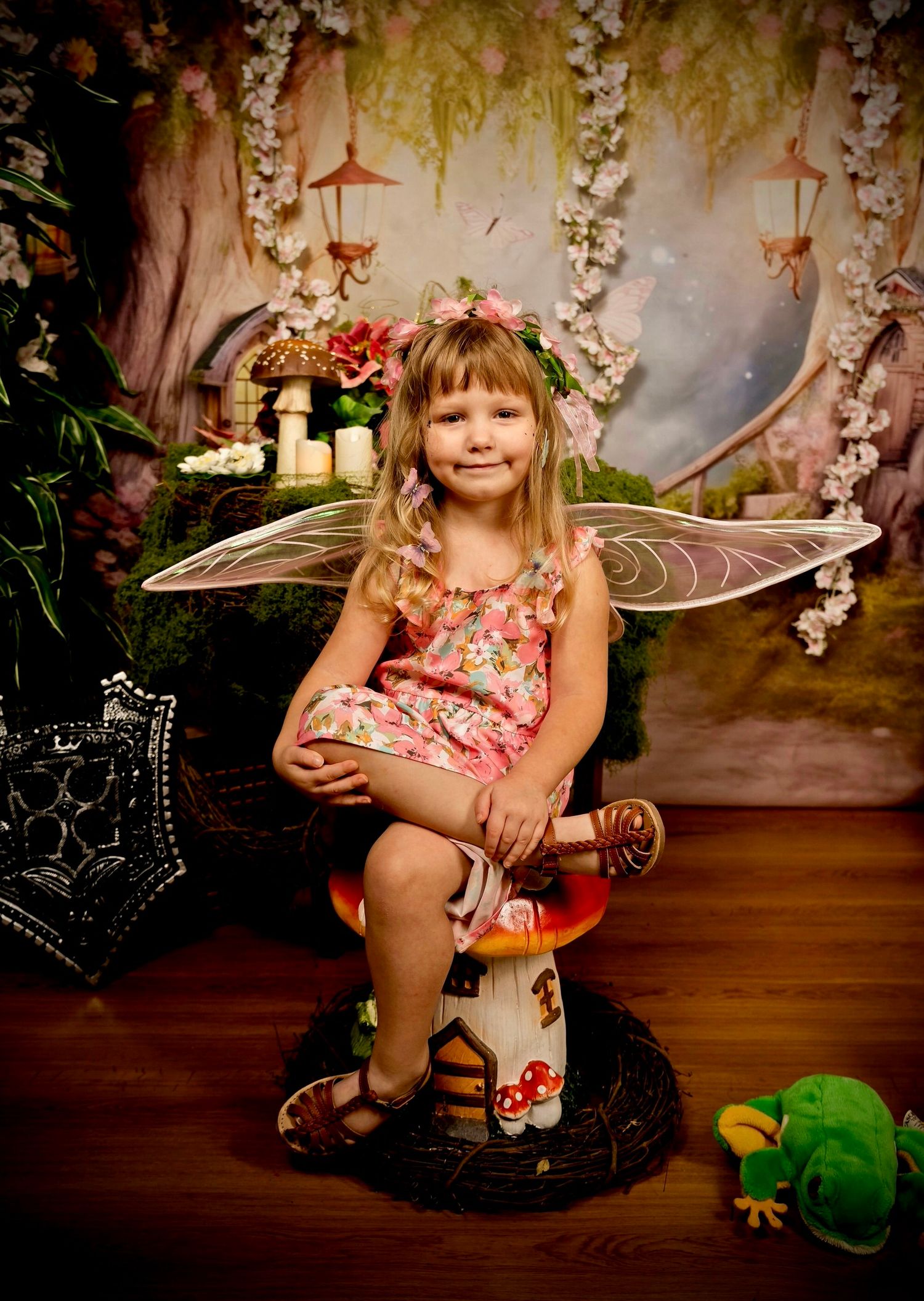 Young girl dressed as a fairy with wings and floral dress sits on mushroom stool in enchanted forest photo backdrop.