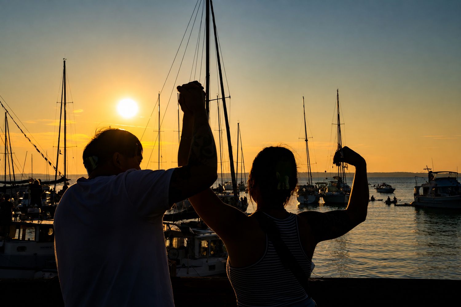 Two silhouettes flexing muscles at sunset by a marina with sailboats in the golden background.