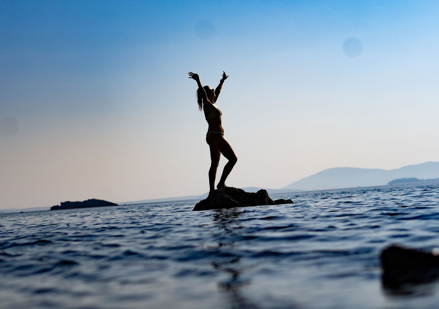 Silhouette of a woman standing on a rock in the ocean with arms raised against a blue sky.