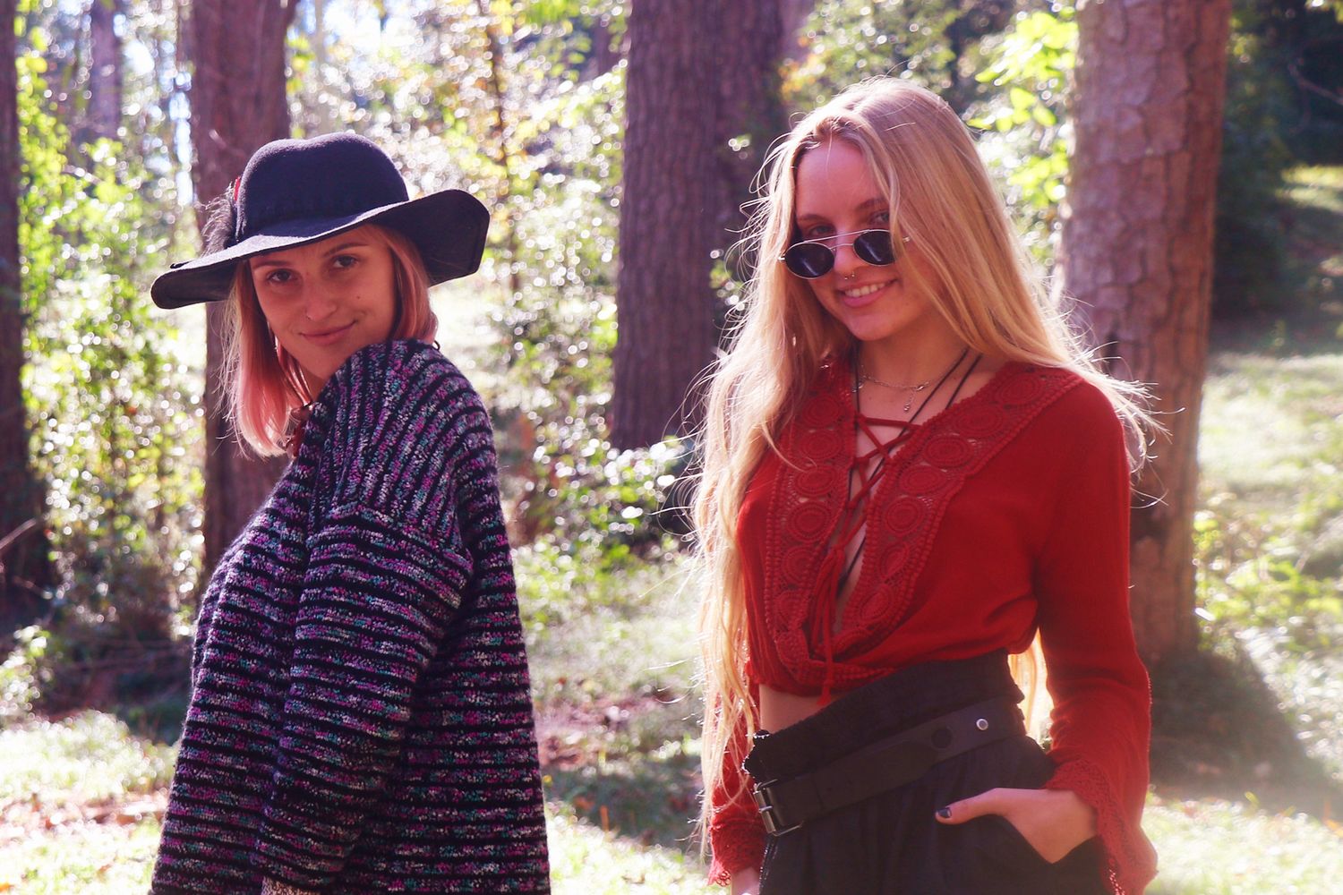 Two young women posing outdoors in a sunlit forest, one wearing a black hat and the other in a red top with sunglasses.