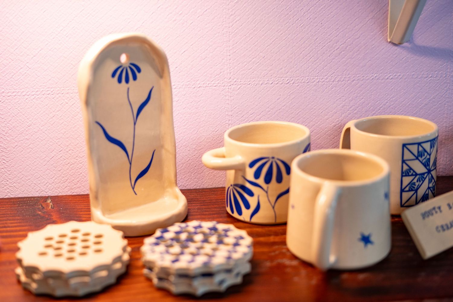 Handcrafted blue and white ceramic mugs, coasters, and a wall vase with floral designs displayed on a wooden shelf.
