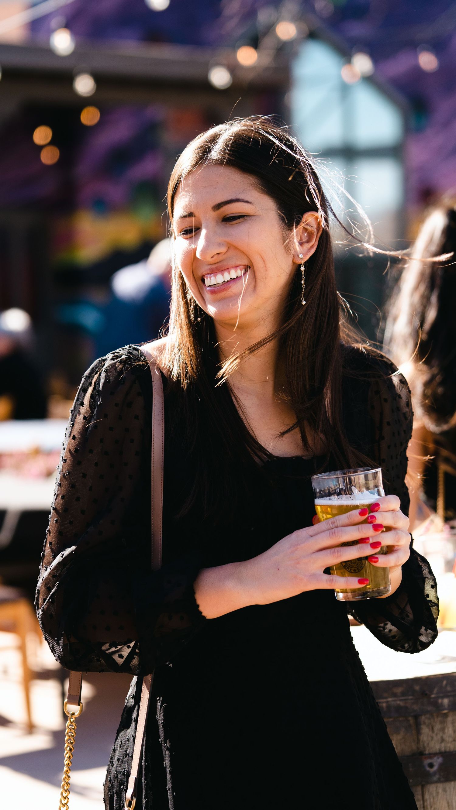 Smiling woman in black lace dress holding a beer glass outdoors at a sunny social event.