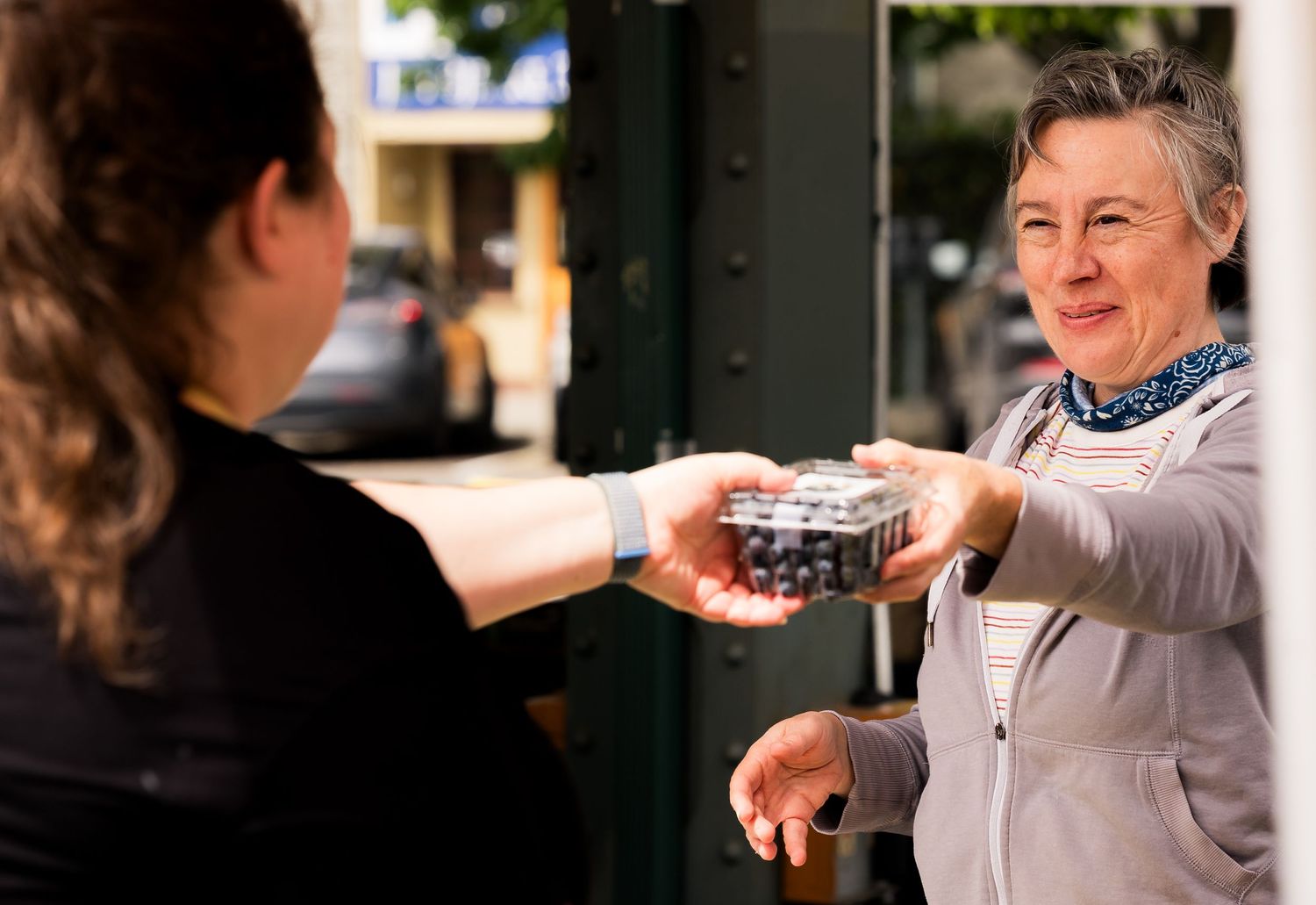 Woman in gray jacket receiving a container of blueberries from a store employee outdoors.