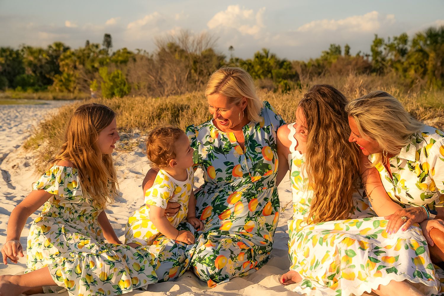 A photo of a family of girls smiling at each other on the beach, wearing patterned dresses.
