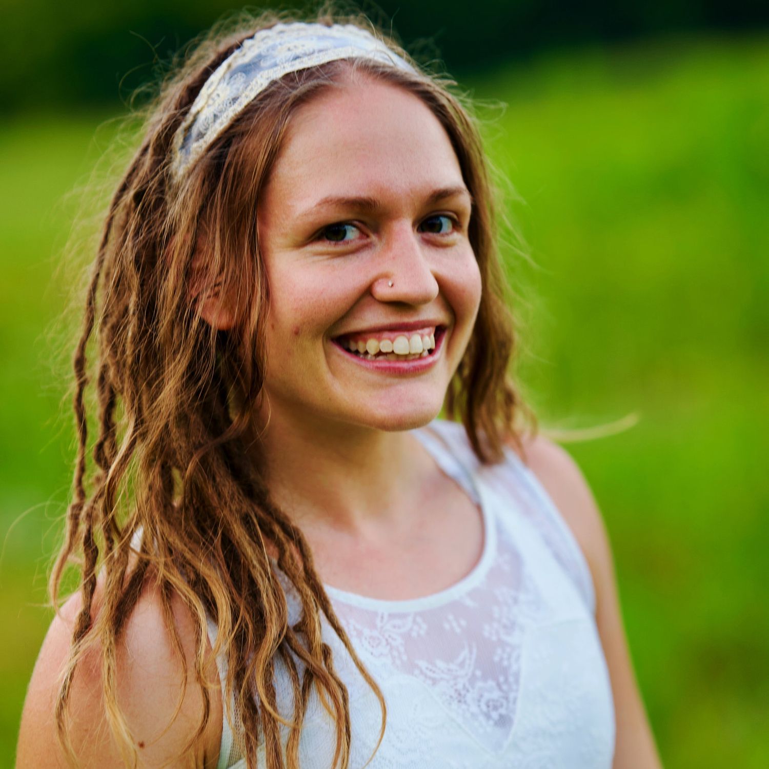 A photo of a woman with dreads smiling at the camera, she is wearing a white tank top and is against a green backdrop.