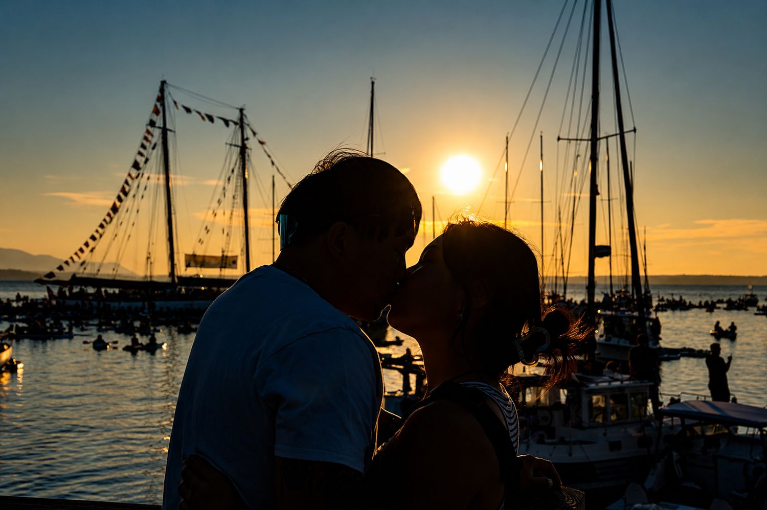 Couple sharing a romantic kiss silhouetted against a golden sunset at a marina with sailboats in the background.