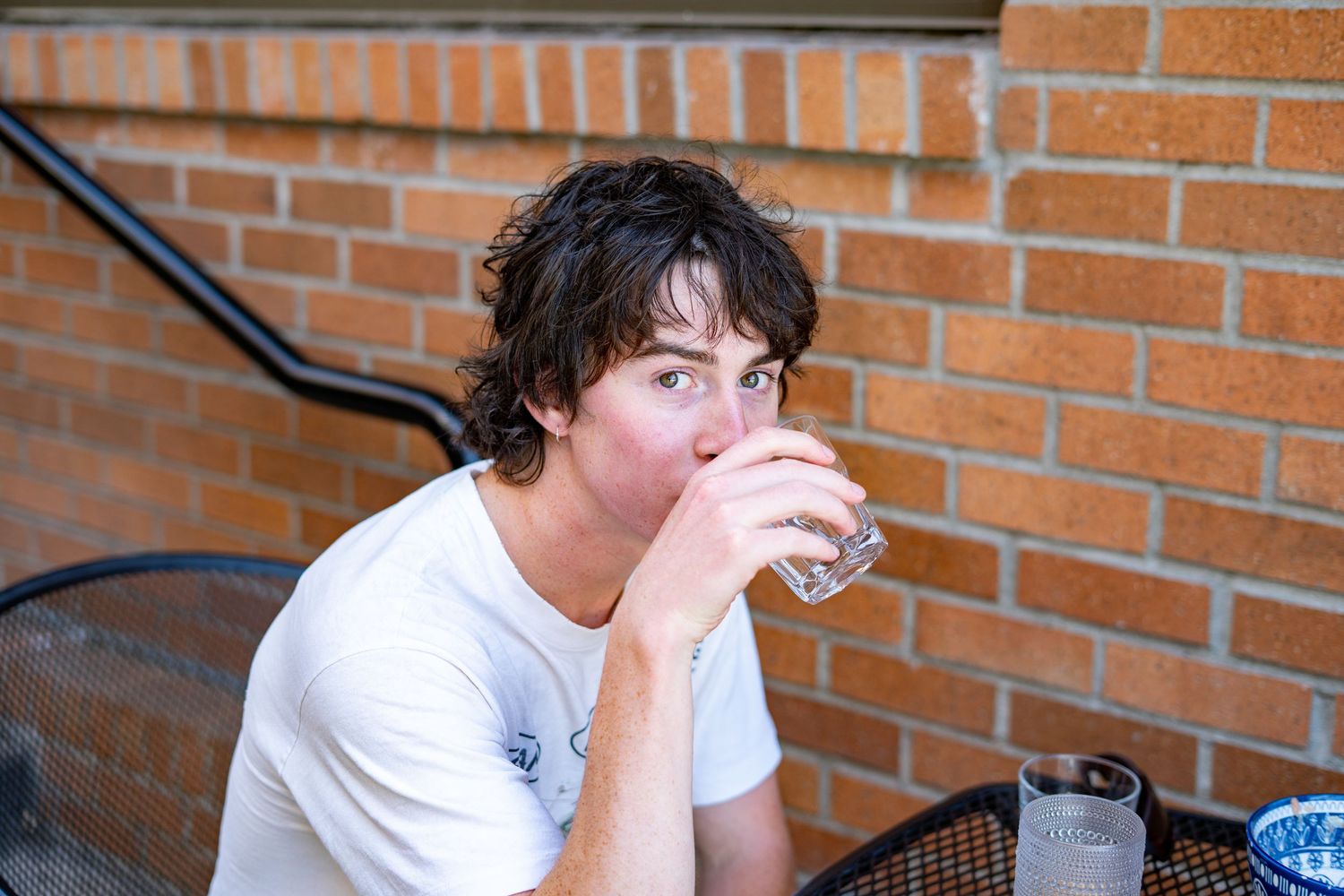 Young man with curly hair drinking water from a glass while sitting at an outdoor patio table near a brick wall.