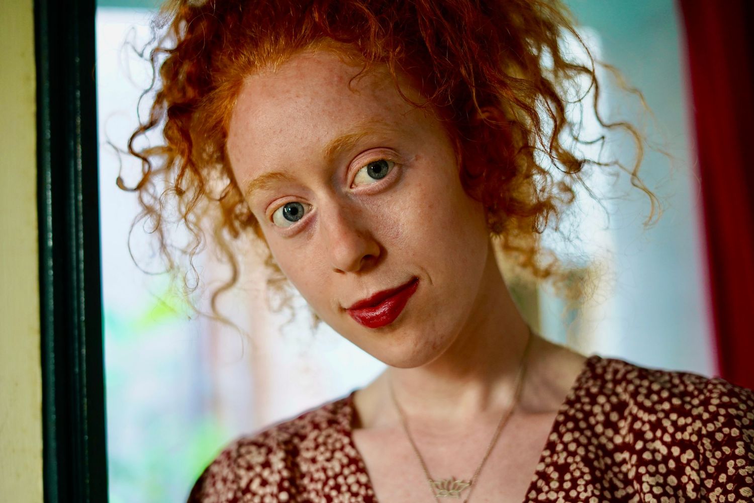 Young woman with curly red hair, blue eyes, and red lips wearing a floral top and delicate necklace.