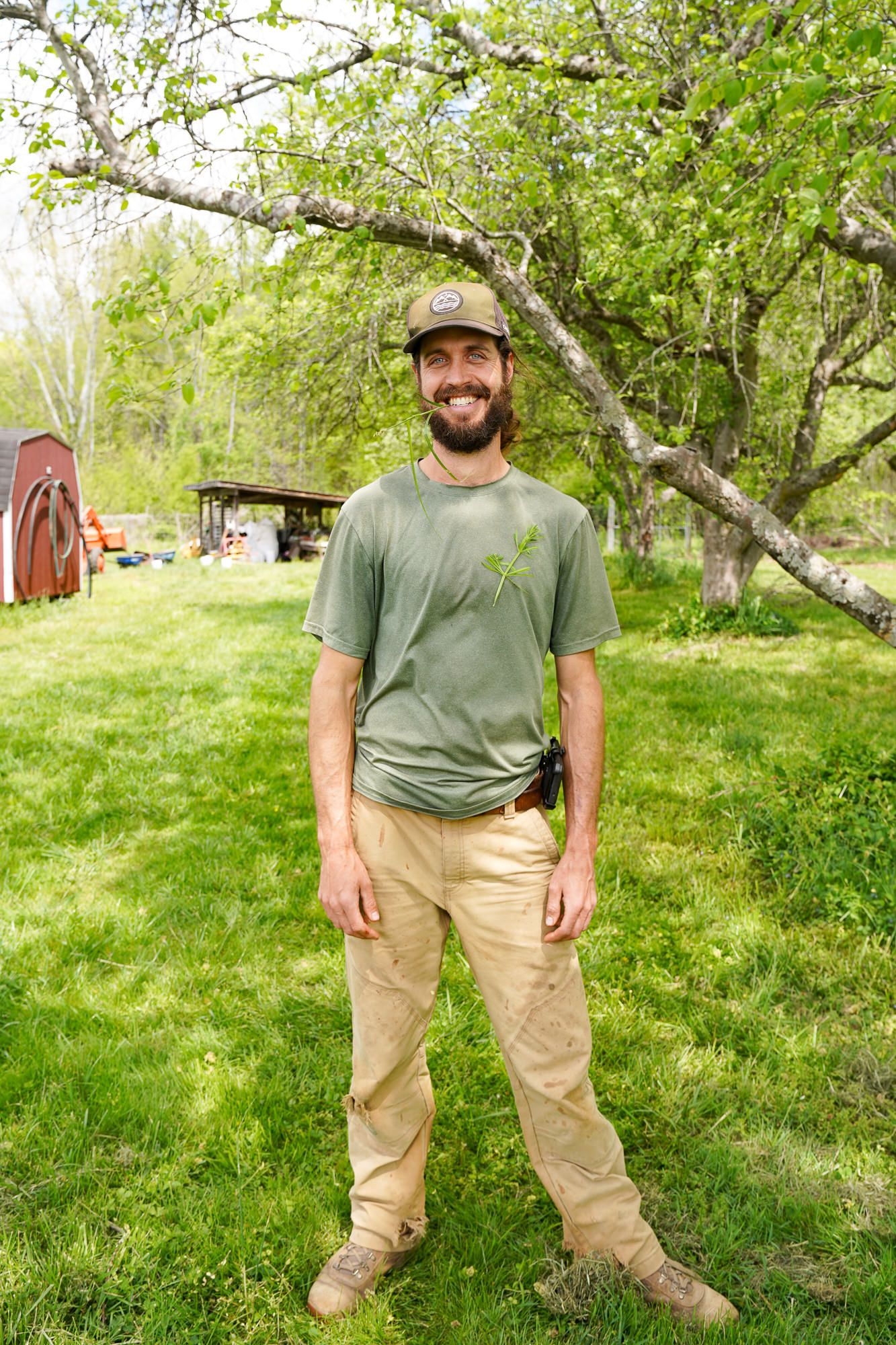 Smiling bearded man in green t-shirt and khaki pants standing outdoors on lush green grass near trees.