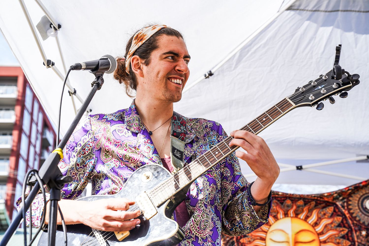 Smiling male guitarist performs outdoors in colorful floral shirt at sunny festival stage event.