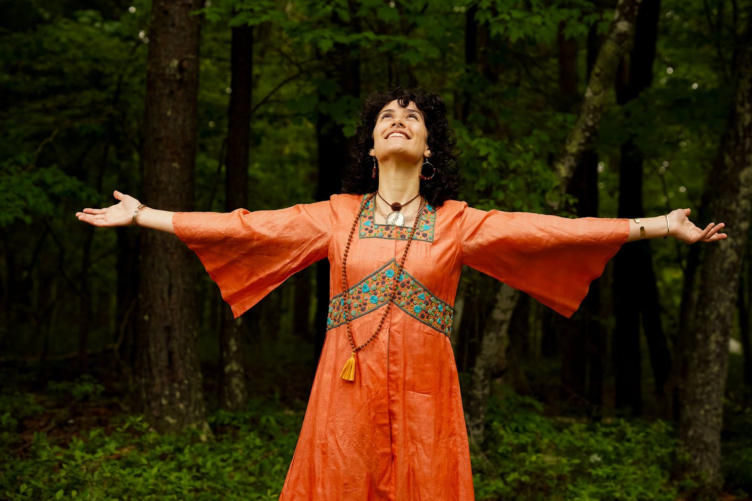 Woman in orange kimono dress with arms outstretched in green forest.