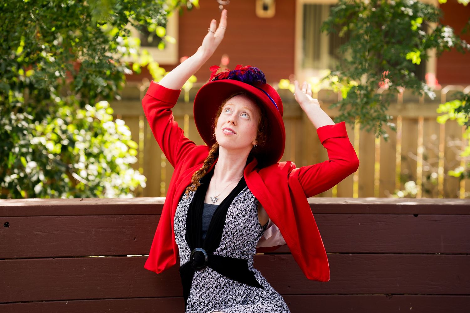 Woman in red cardigan and hat poses gracefully on a bench surrounded by lush green trees.
