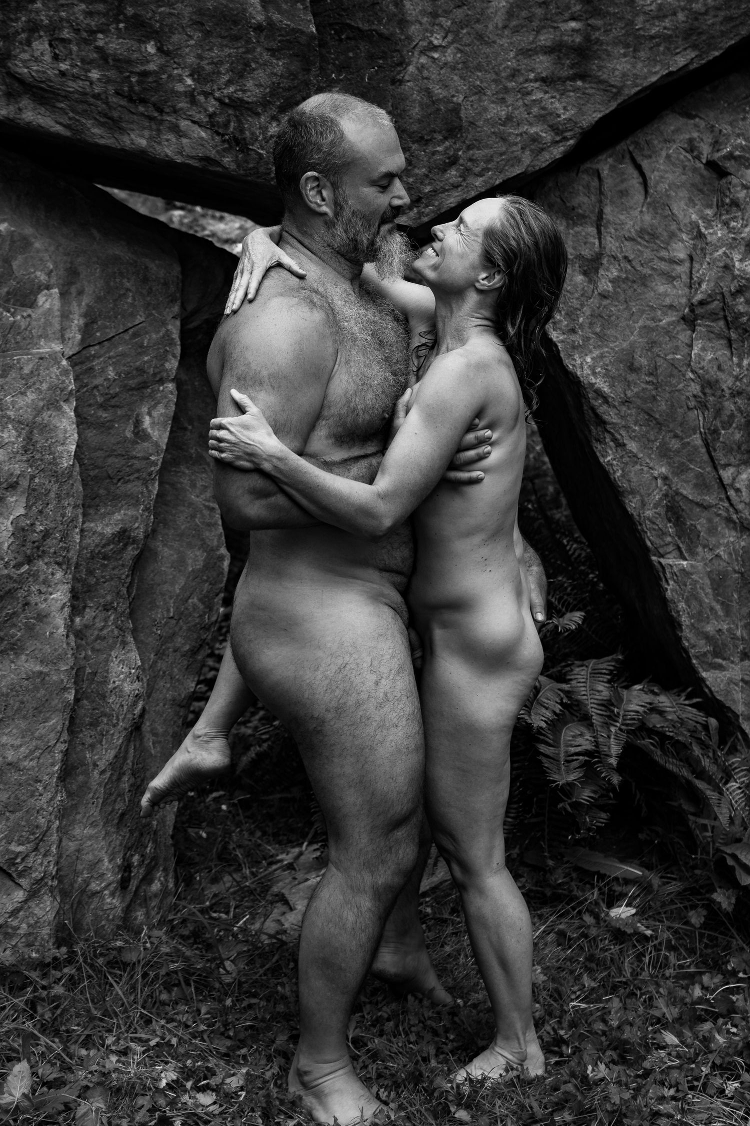 Black and white fine art nude photograph of a couple embracing outdoors against rocky natural background.