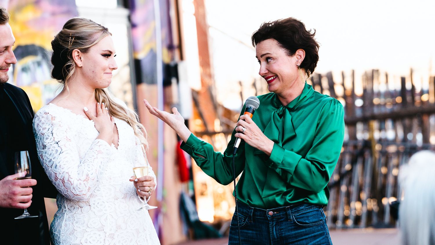 Woman in green shirt holds microphone interviewing bride in white lace dress at outdoor wedding reception.