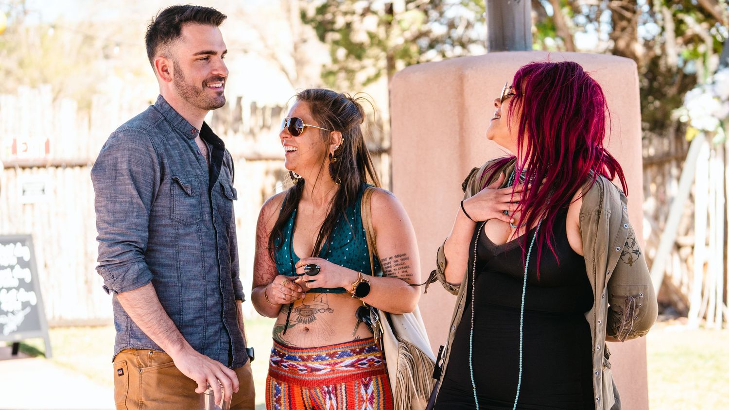 Three friends chatting outdoors at a festival, woman in bohemian crop top with tattoos between man and red-haired woman.
