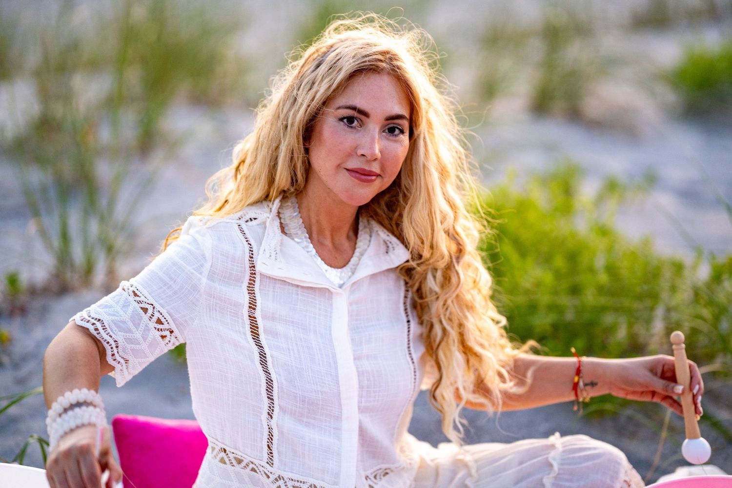A photo of a woman with blonde flowy hair playing singing bowls on the beach, she is wearing a white button up top.