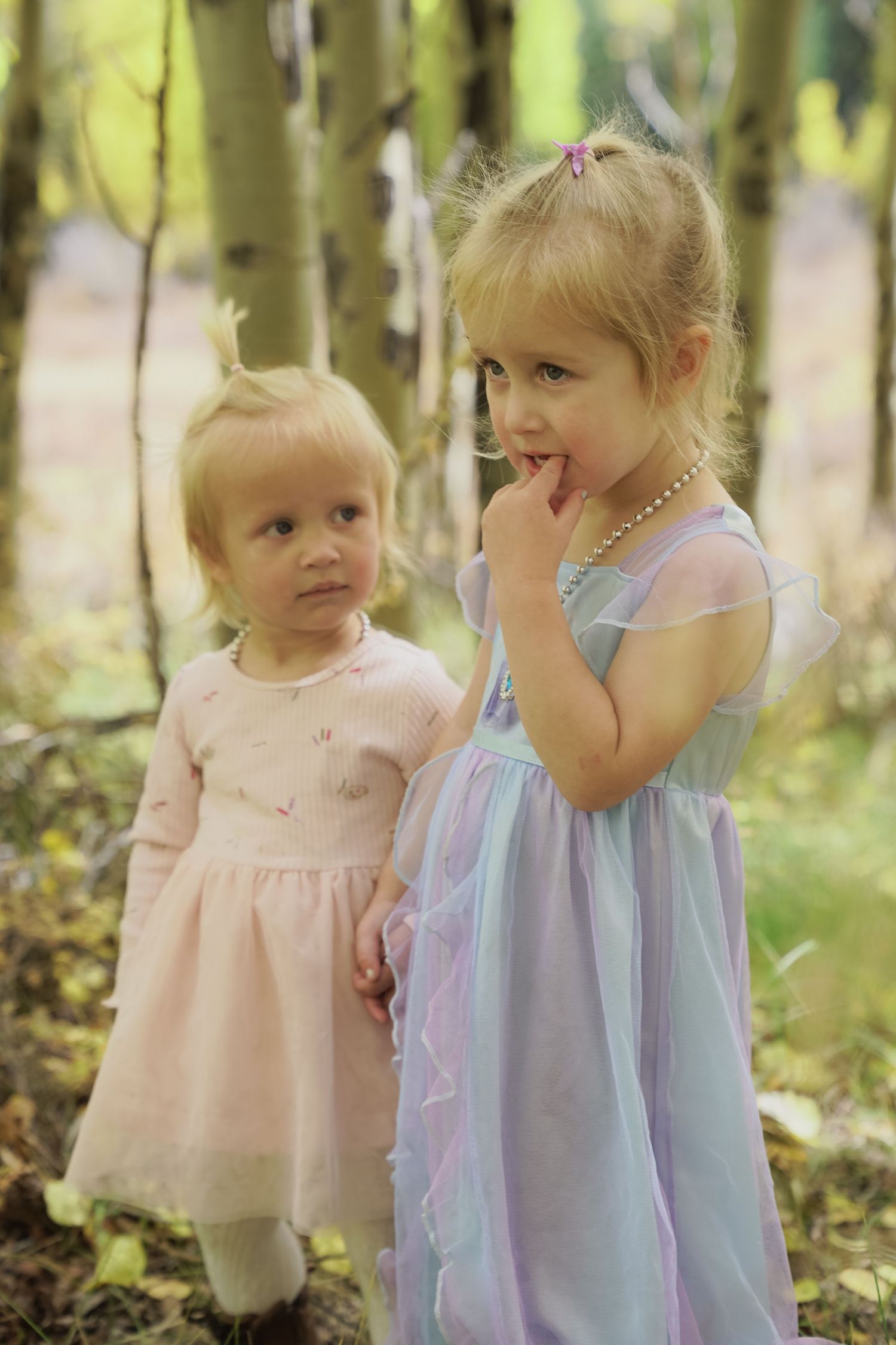 Two young children in pastel dresses explore a sunlit forest setting together.